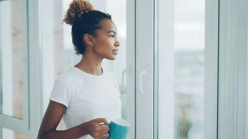Une jeune femme en tee-shirt blanc, une tasse à la main, regarde par la fenêtre, l'air apaisé.