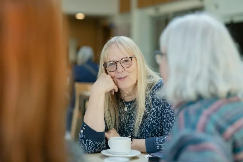 Une femme aux cheveux blonds et aux lunettes écoute, la main contre la joue, une amie aux cheveux gris attablée devant un café.