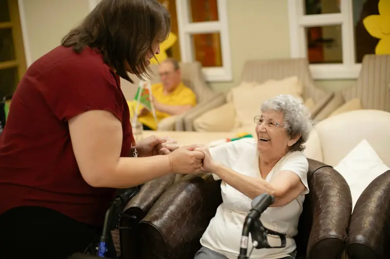 Une intervenante en polo bordeaux tient les mains d'une dame aux cheveux blancs assise dans un fauteuil, qui sourit.