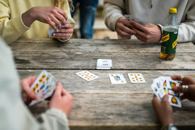 Vue de dessus d'une partie de cartes sur une table en bois : des mains tiennent leur jeu, d'autres cartes sont posées au centre.