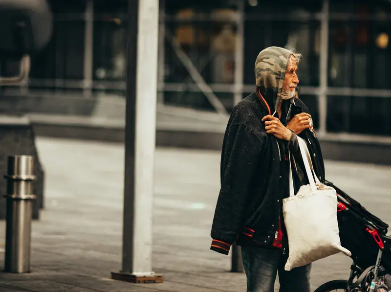 Une personne âgée en manteau sombre, un sac en toile à l'épaule, tire un petit chariot sur un trottoir.