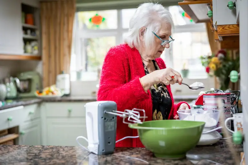Une dame aux cheveux blancs, gilet rouge, prépare un plat dans sa cuisine, un saladier vert devant elle.
