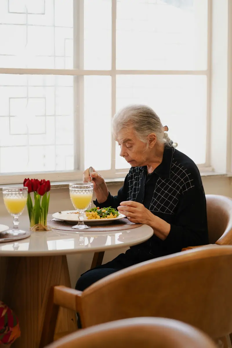 Un monsieur aux cheveux gris déjeune à une table ronde près d'une grande fenêtre, un bouquet de tulipes rouges devant lui.