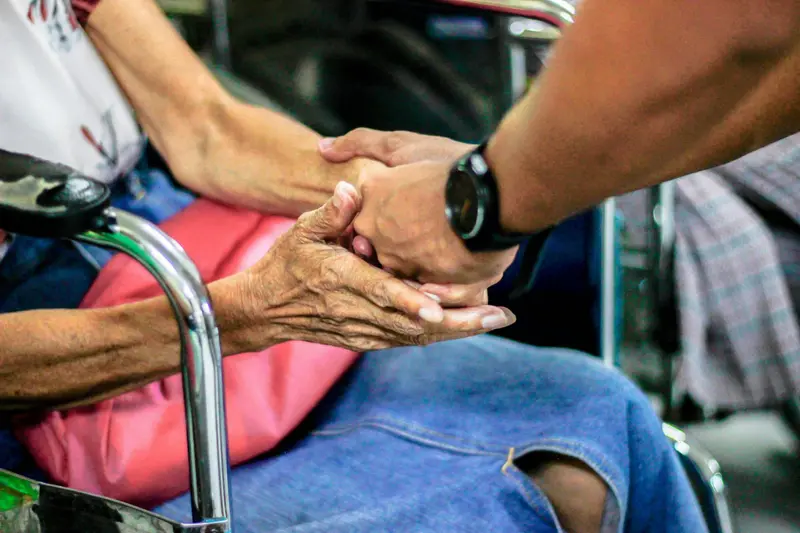 Gros plan sur deux mains qui se tiennent : celle d'une personne âgée, posée sur son genou, et celle d'un homme portant une montre.