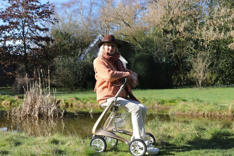 Une dame en chapeau et manteau rose se promène au bord d'un étang, appuyée sur son déambulateur.