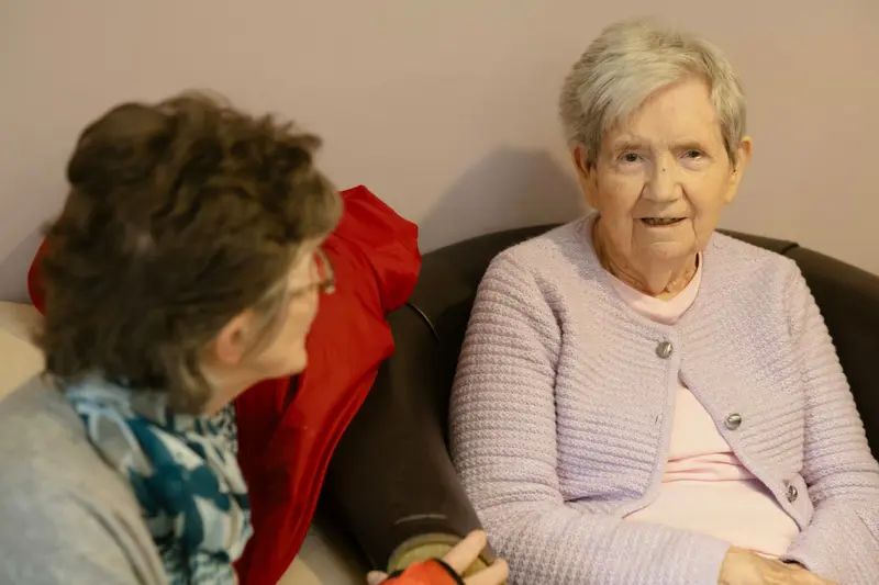 Deux dames âgées bavardent dans un salon ; l'une, en gilet mauve, sourit.